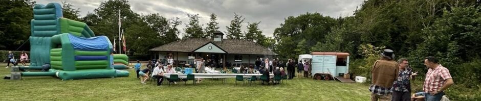 A village green wit a bouncy castle a few tables, a horsebox bar and people drinking and talking